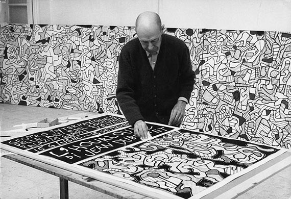 Abbildung zu: Jean Dubuffet in seinem Atelier in Vence, 1964, mit dem Plakat der „L'Hourloupe“-Ausstellung, Palazzo Grassi, Venedig, Foto: Max Loreau. © Archives Fondation Dubuffet, Paris / © VG Bild-Kunst, Bonn 2025